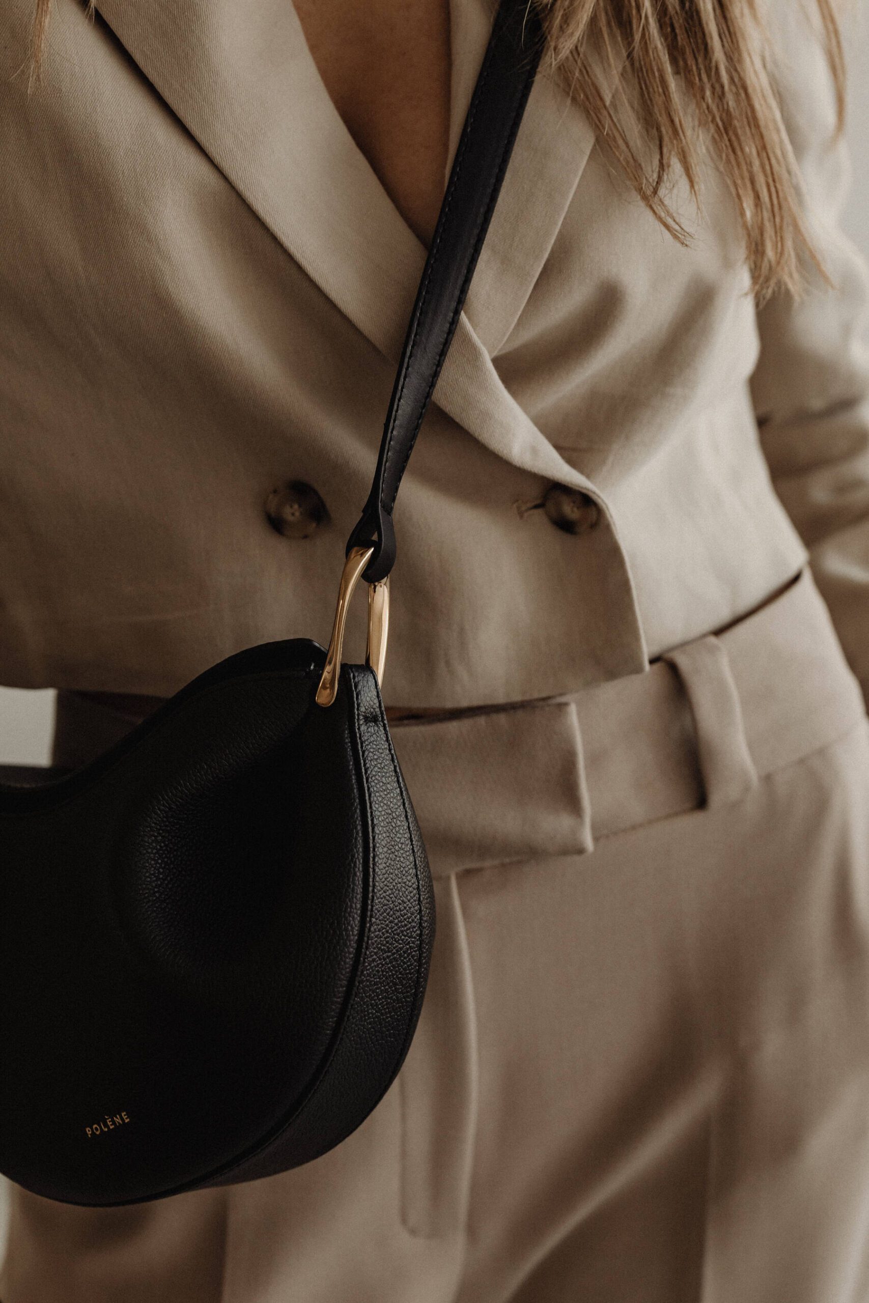 A person wearing a beige suit with long sleeves and a deep neckline holds the elegant Milla small black leather crossbody bag with gold hardware. The bag, perfect for carrying your portfolio essentials, rests at their waist. Inner North Bookkeeping