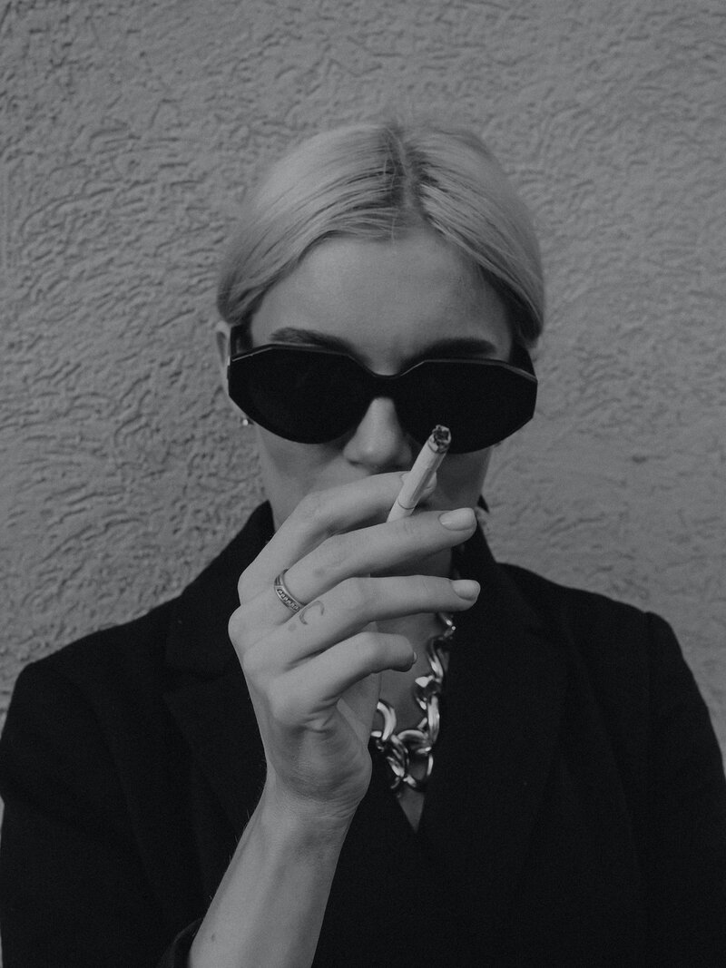 A person wearing dark sunglasses and a chunky necklace holds a cigarette towards the camera, exuding an enigmatic presence perfect for a Portfolio feature. They have light hair and are dressed in dark clothing, standing against a textured wall. The black and white photo captures the striking style reminiscent of Milla’s iconic looks. Inner North Bookkeeping