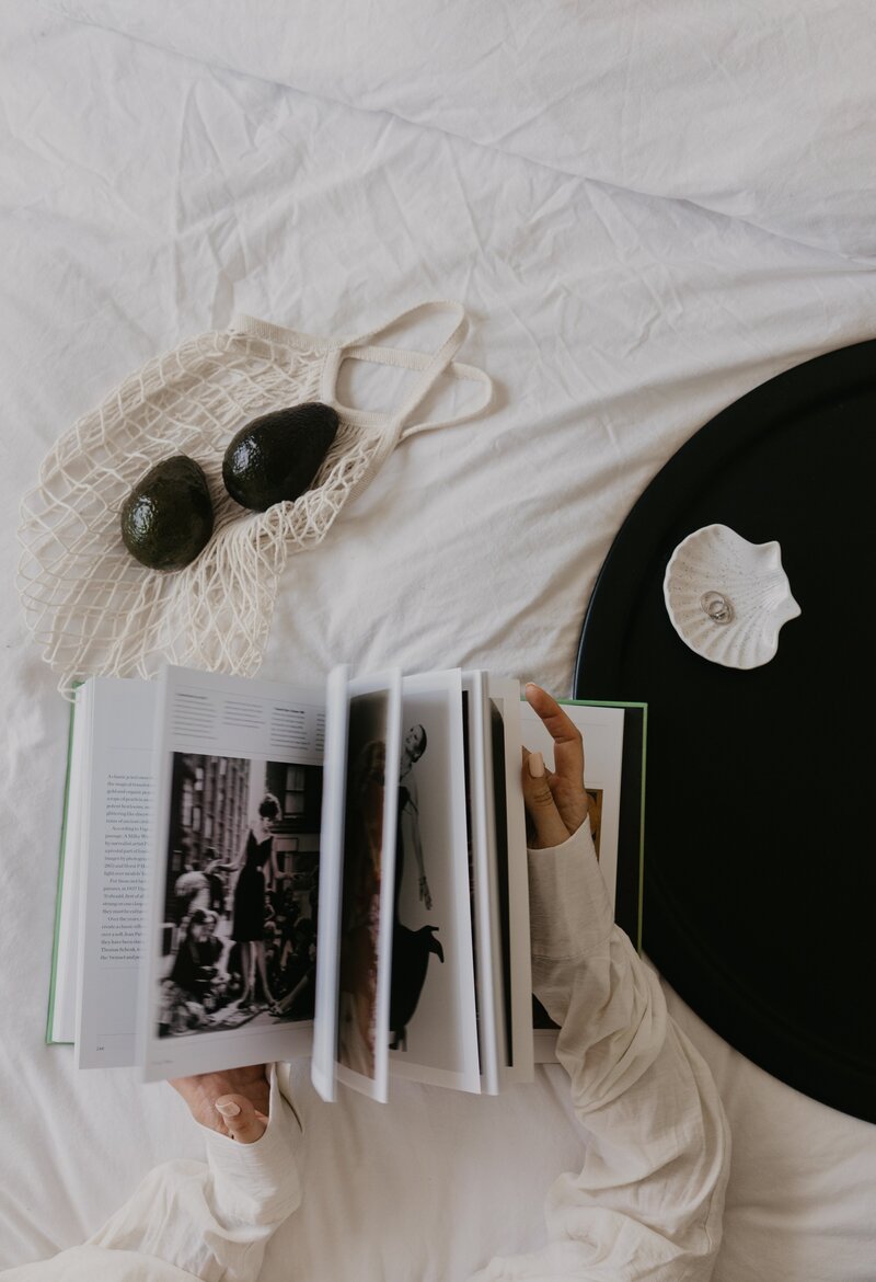 A person reads a photo book on white bedding next to a black tray with a shell dish and a reusable bag containing two avocados. Inner North Bookkeeping