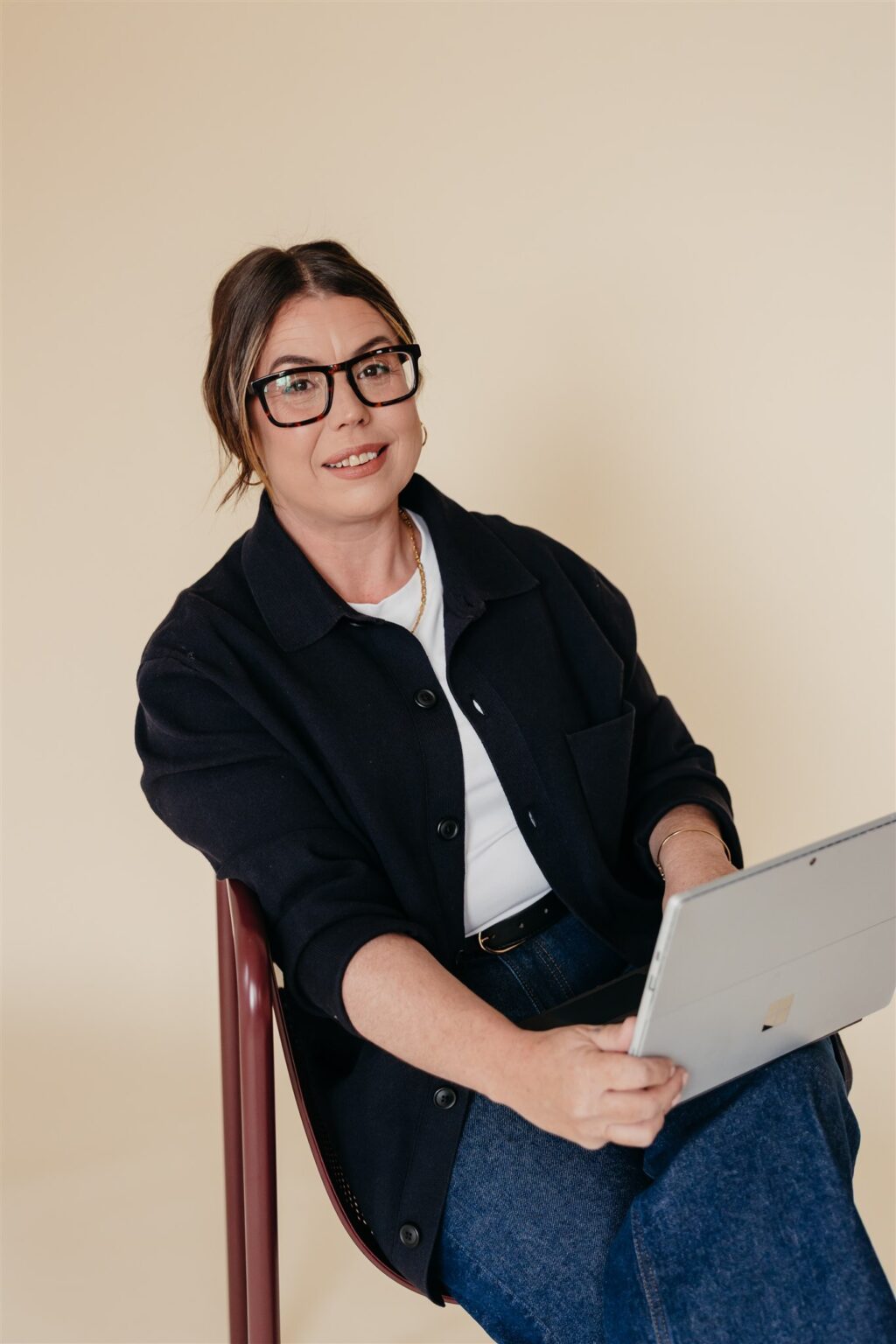 A woman with glasses, wearing a dark jacket and jeans, sits on a chair holding a tablet and smiling at the camera against a beige background. Inner North Bookkeeping