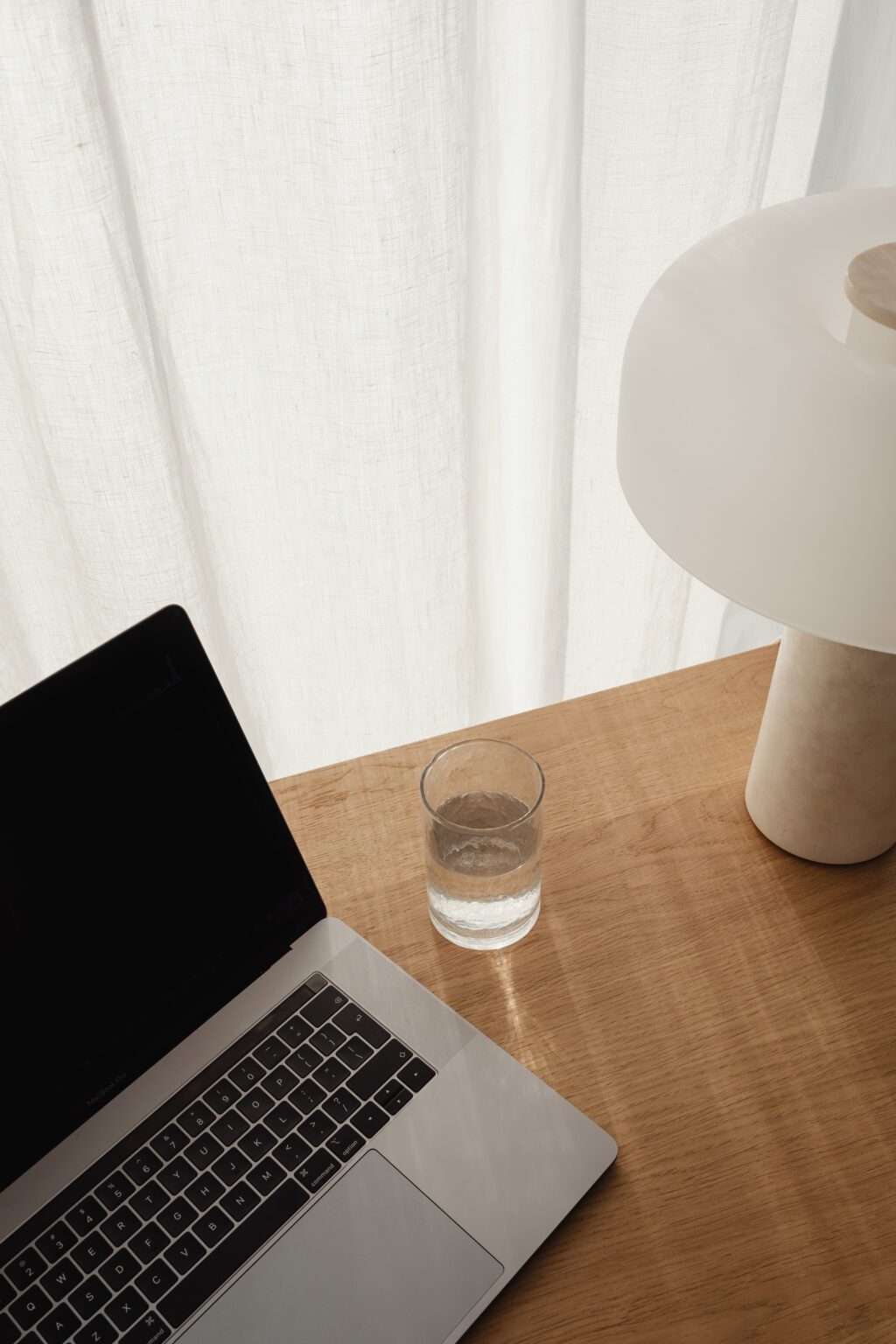 A glass of water, a closed laptop, and a modern white lamp sit on a wooden desk—an inviting workspace for bookkeepers Melbourne relies on—with sunlight filtering through a sheer white curtain in the background. Inner North Bookkeeping