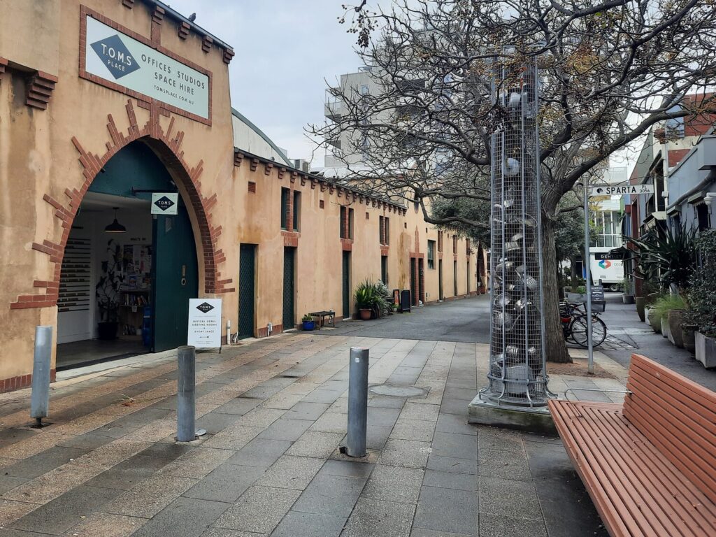 A quiet pedestrian street with a tan building labeled “TOMS OFFICE STUDIOS SPACE HIRE,” benches, a leafless tree, and a bicycle in the background. The scene appears calm and overcast—an ideal spot for a bookkeeper for small business to find inspiration or take a peaceful break between tasks. Inner North Bookkeeping