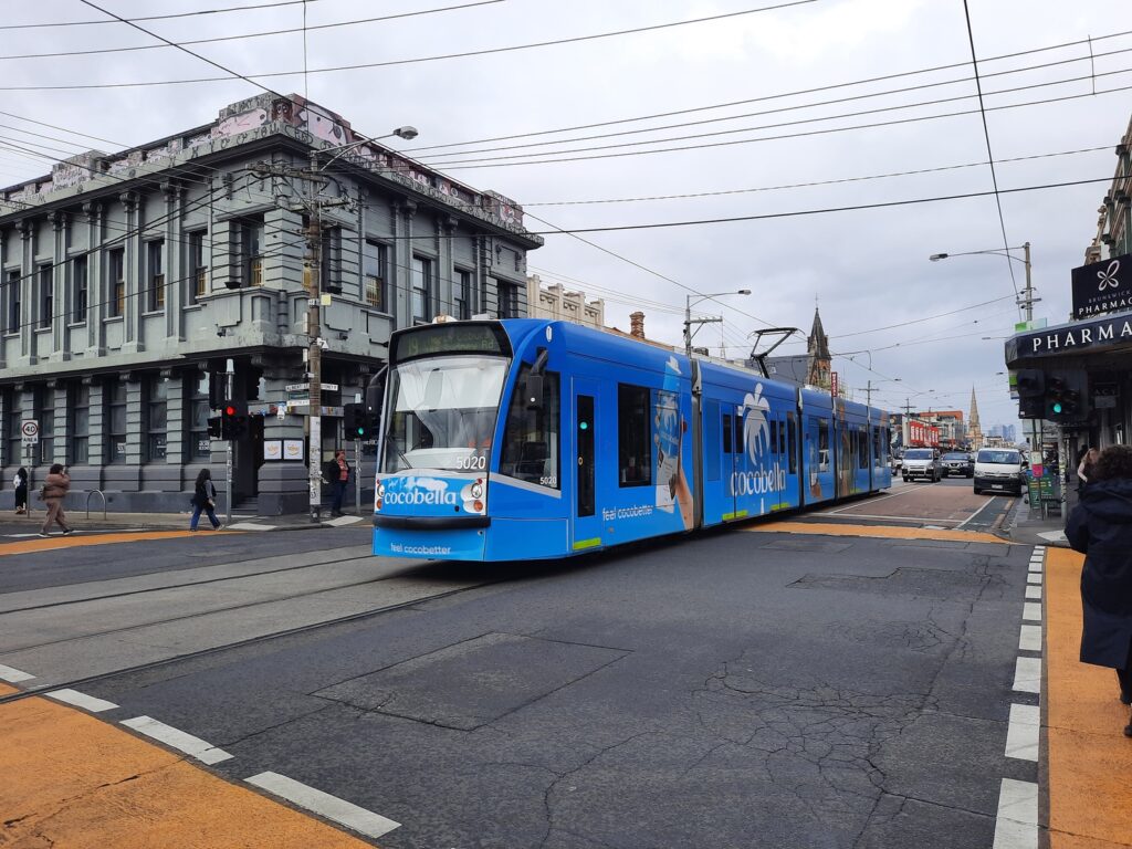 A blue tram with advertising travels through a city intersection, passing old buildings. Pedestrians walk on the sidewalks and cross the street under a cloudy sky. Inner North Bookkeeping